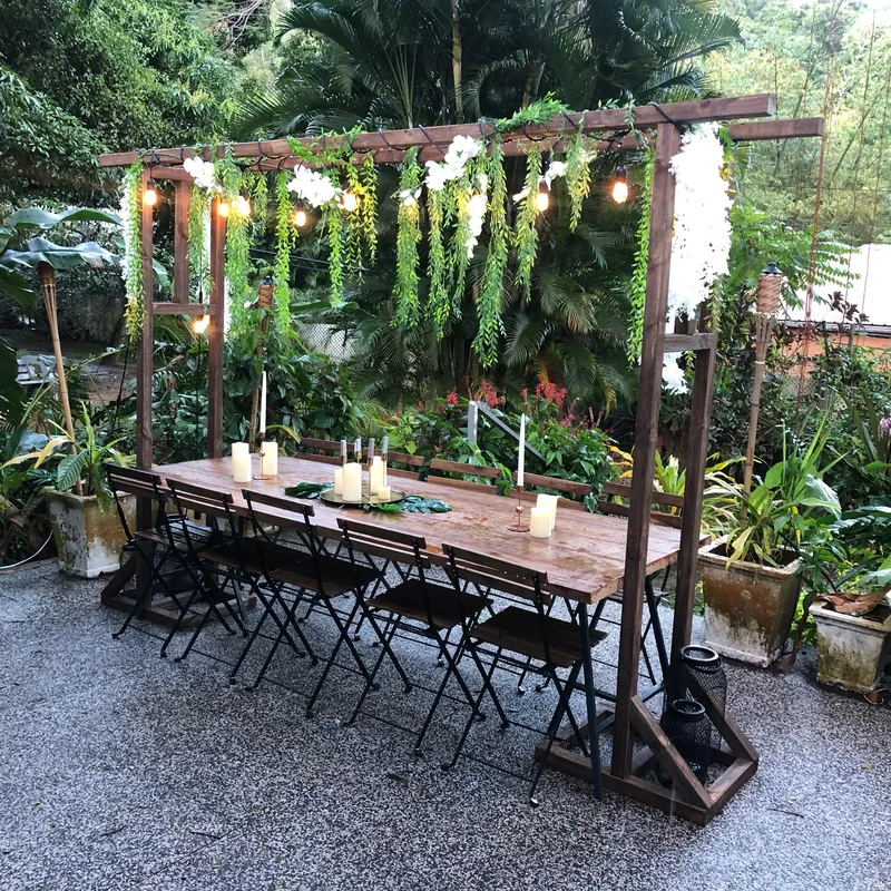 The Pergola Arch & Table outdoors: a wooden table and black chairs under a frame with greenery, white flowers and lights