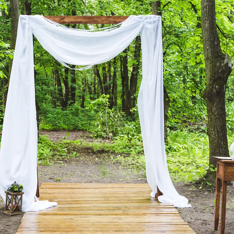 The Ceremony Arch draped with white fabric on a wooden walkway in a green forest, with a small table and a lantern beside it