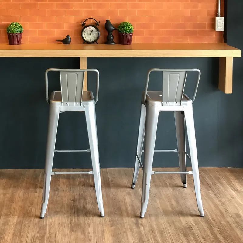 Two Industrial Bar Stools at a wooden counter against an orange brick-patterned wall with a clock, plants and bird figurines