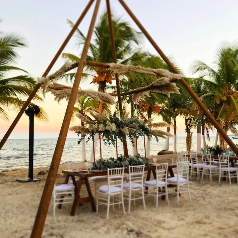 Naked Tipi with a bamboo canopy above wooden tables and white chairs on a beach near palm trees and the sea at sunset