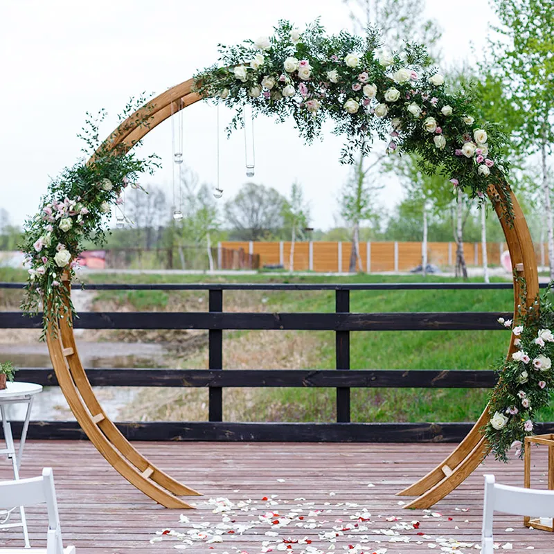 Oval Arch decorated with white and pink flowers and greenery on an outdoor wooden platform, with chairs and petals in front