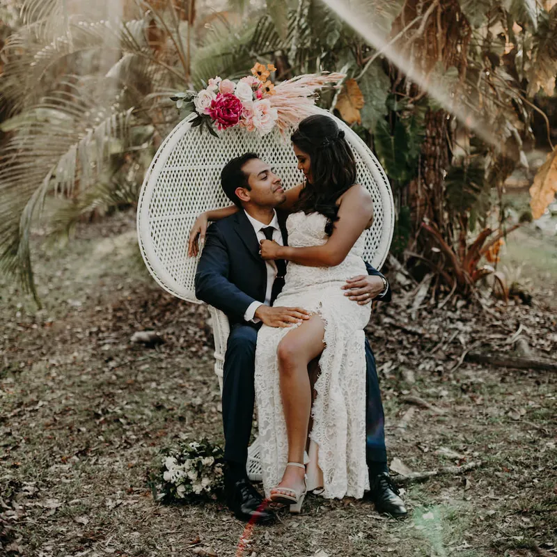 A bride in a white lace dress sits on a groom's lap on a Peacock Chair decorated with flowers, outdoors in a wooded area