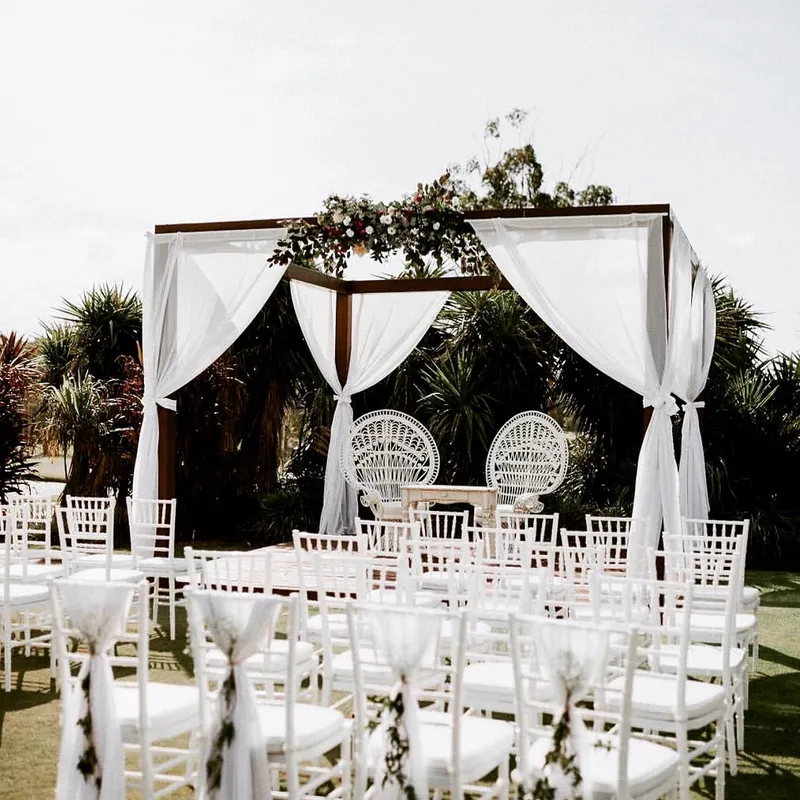 The Pergola Arch with drapes at an outdoor wedding set-up, with white chairs, two decorative chairs beneath it and greenery