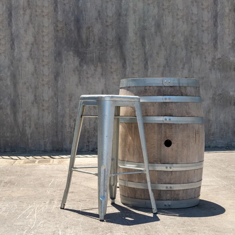 Rum Barrel, a wooden barrel with metal bands, beside a metal stool on concrete against a grey textured wall