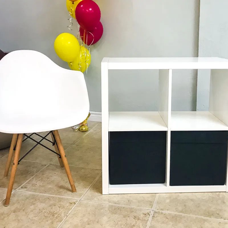 Simple Storage Table with two black bins beside a white chair with wooden legs, with red and yellow balloons behind