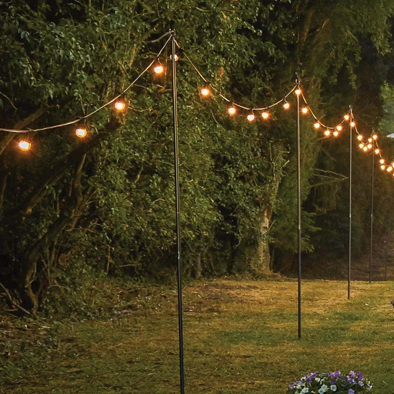 String Light Poles with string lights hanging between them over a grassy area bordered by dense trees, flowers in the corner