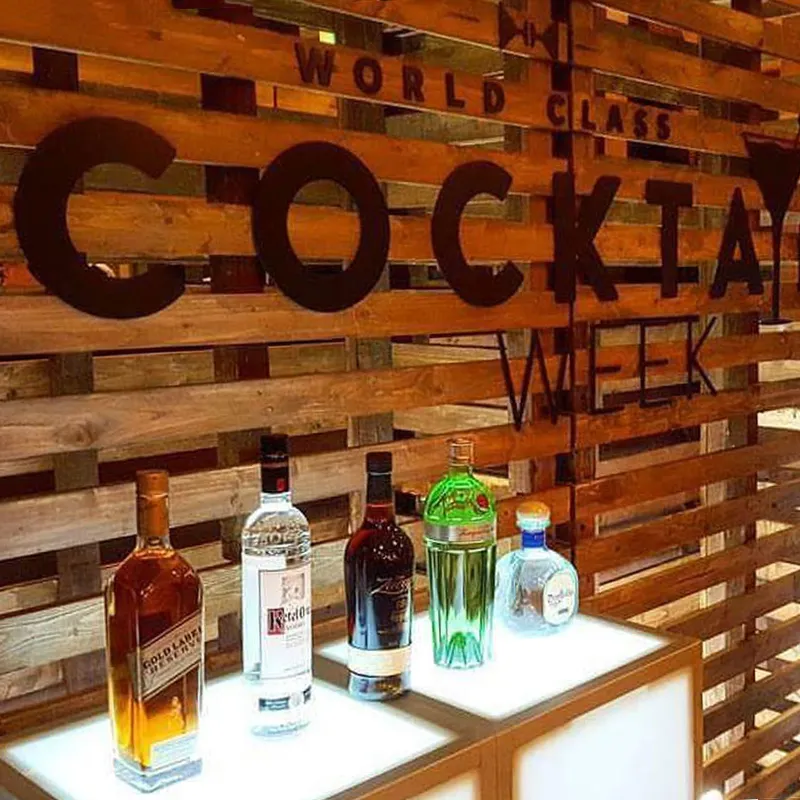 A Pallet Wall Backdrop with “World Class Cocktail Week” in large black letters, and five liquor bottles on a lit counter