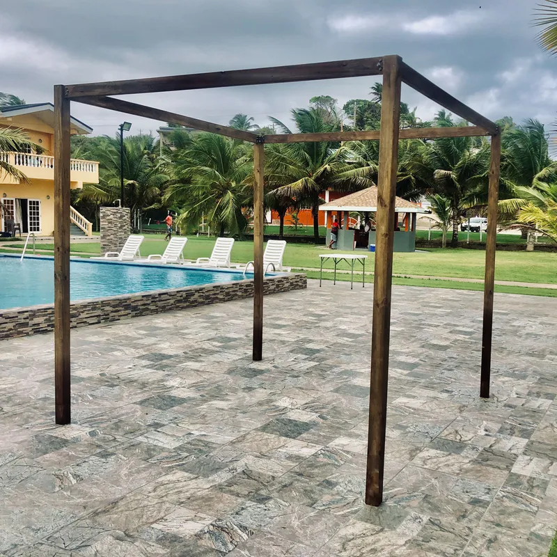 A Pergola Arch standing on a tiled poolside patio with loungers, a swimming pool and palm trees behind it under a cloudy sky