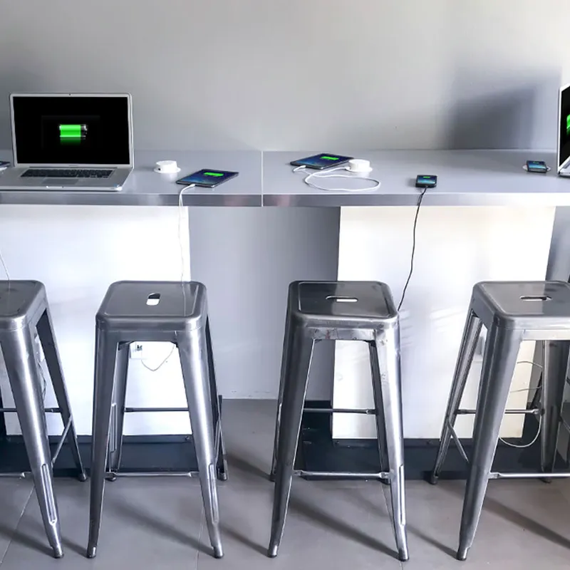 Four Wireless Charging Breakout Tables (4ft) with metal stools, each charging laptops and phones that show charging screens