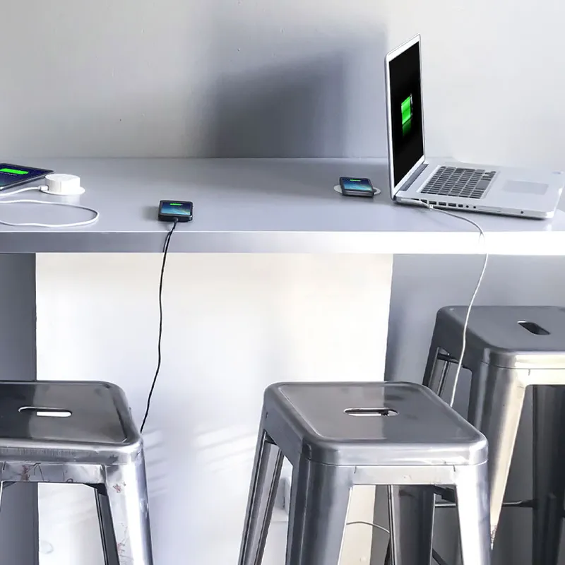 A laptop, phone and tablet charging on the Wireless Charging Breakout Table, with metal stools underneath, in a modern space
