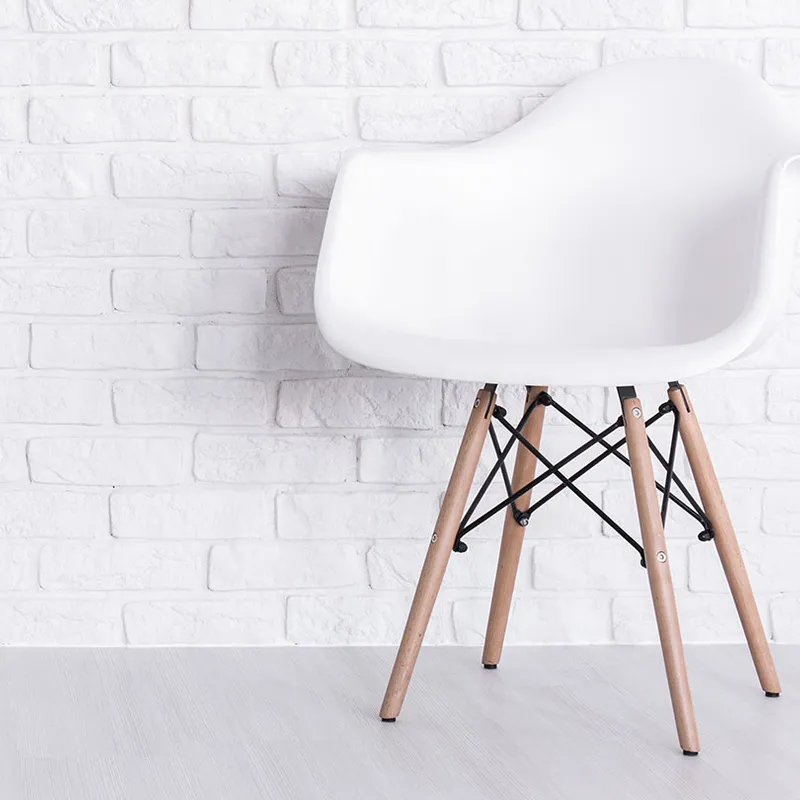 Eames Armchair, a modern white plastic chair with wooden legs, against a white brick wall on a light-coloured floor