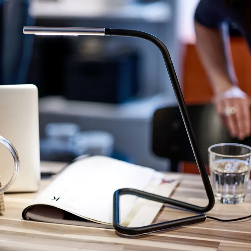 Flex Lamp, a black LED desk lamp with an adjustable head, on a wooden table beside an open magazine and a glass of water