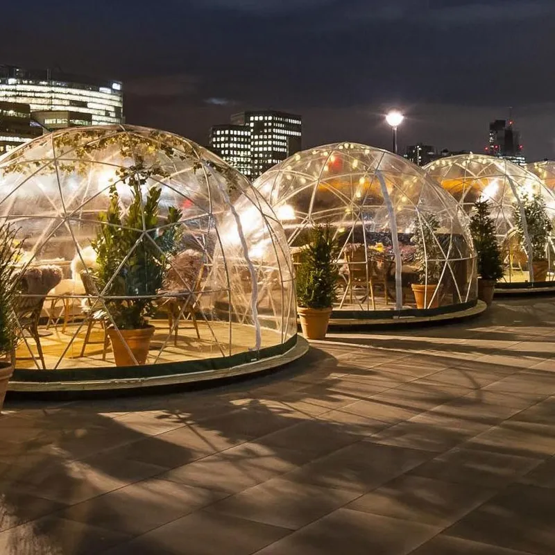 A row of Geodesic Air Conditioning Domes with tables and chairs on an outdoor terrace at night, with potted plants