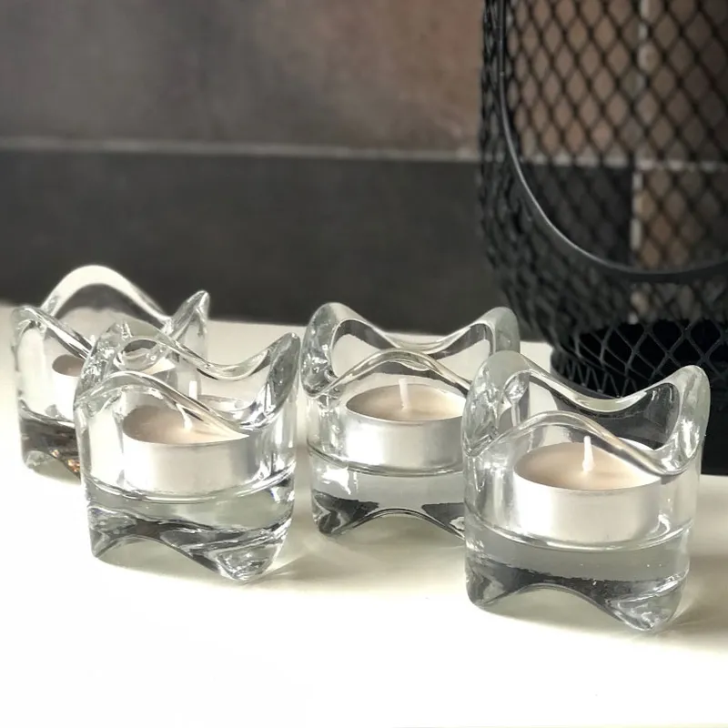 Four Wave Tealight glass candle holders with white candles, arranged on a white surface beside a black mesh décor piece