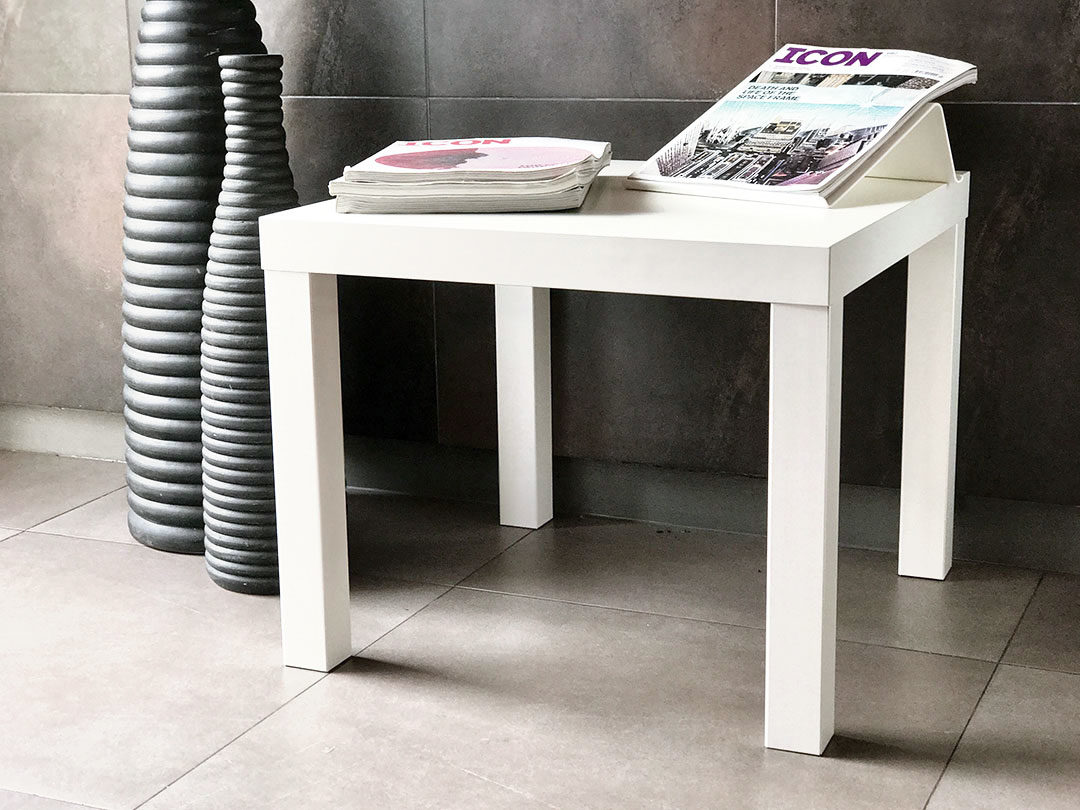 A simple side table from Trinidad Event Furniture, white and rectangular, holds stacked magazines and sits on a tiled floor beside a tall, textured black vase. - Perched Event Rentals, Trinidad