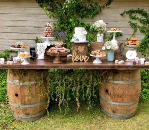 The Rum Barrel Bar from Trinidad Event Rentals displays pastries, a white cake, and flowers on wooden barrels outdoors, accented with greenery and a charming “love” sign for a rustic dessert table setup. - Perched Event Rentals, Trinidad