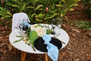 A small round table set for two outdoors, featuring black plates, blue serviettes, glassware, candles, gold cutlery, and a centrepiece with white flowers and green foliage from Trinidad Event Rentals. - Perched Event Rentals, Trinidad